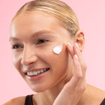 Woman applying cream to her face against a pink background