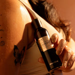 Person holding a skincare bottle with a tattooed arm in the background