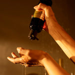 Hand holding a bottle over another hand with water dripping, against a warm-toned background