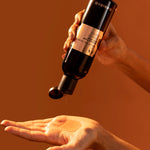 Hand holding a black bottle with a dropper, dispensing liquid onto another hand against an orange background