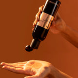 Hand holding a black bottle with a dropper, dispensing liquid onto another hand against an orange background