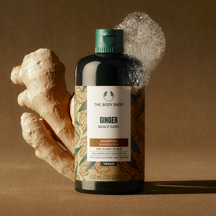 Bottle of The Body Shop Ginger Scalp Care Shampoo with a piece of ginger on a brown background