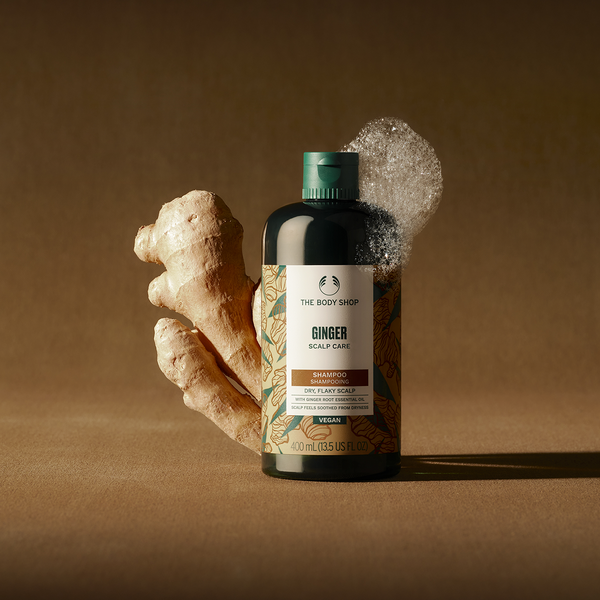 Bottle of The Body Shop Ginger Scalp Care Shampoo with a piece of ginger on a brown background