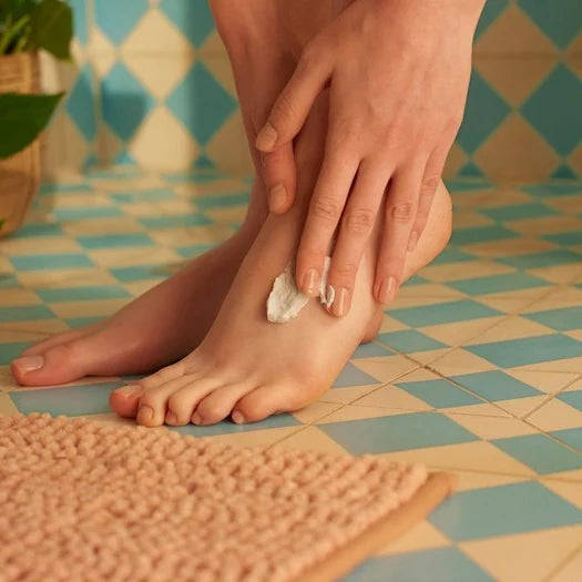 Person applying cream to their foot on a tiled bathroom floor.