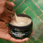 Hand holding a jar of The Body Shop Jamaican Black Castor Oil mask with a green tiled background.