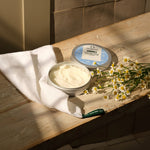 Two containers of body butter on a wooden surface with a white cloth and flowers.