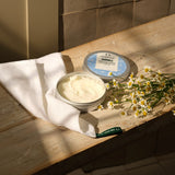 Two containers of body butter on a wooden surface with a white cloth and flowers.