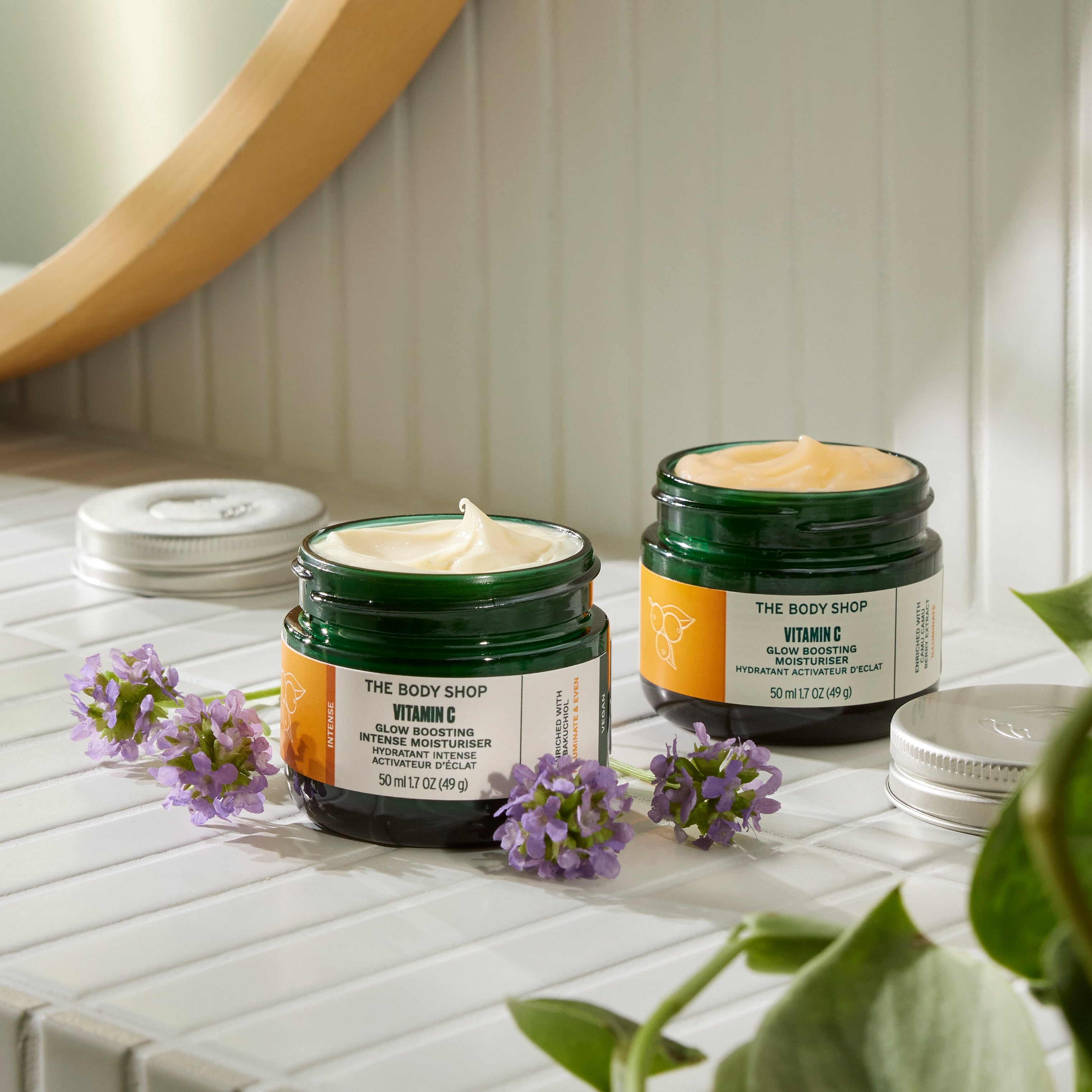 Two jars of The Body Shop product on a white surface with a plant in the foreground.