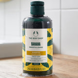 Bottle of The Body Shop Banana Smooth & Shine shampoo on a wooden surface with a white tiled wall background.