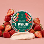 A round tin of The Body Shop Strawberry Body Butter is centered on a pink background, surrounded by fresh sliced strawberries and a dollop of pale cream-colored body butter. The tin label is green and red with the brand name 'THE BODY SHOP' and 'STRAWBERRY' printed prominently.