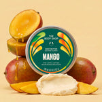 A round tin of The Body Shop Mango Body Butter is centered in front of stacked mangoes and a scoop of white cream on a warm beige background.