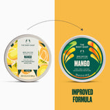 Two round tubs of The Body Shop Mango Body Butter are shown side by side on a split white and mustard yellow background, with a dotted arrow above indicating an improved formula. The left tub has a white lid with yellow citrus illustrations, and the right tub has a dark green lid with orange and yellow abstract leaf-like graphics.