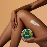A person holds a round tub of The Body Shop Mango Body Butter while applying a thick white cream to their leg against a warm beige background.