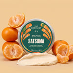 A round tin of The Body Shop Satsuma body butter is centered on a warm beige background, surrounded by whole and sliced oranges, with a smear of creamy off-white body butter in front. The label is dark green with orange and white graphics and text.