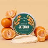 A round tin of The Body Shop Satsuma body butter is centered on a warm beige background, surrounded by whole and sliced oranges, with a smear of creamy off-white body butter in front. The label is dark green with orange and white graphics and text.