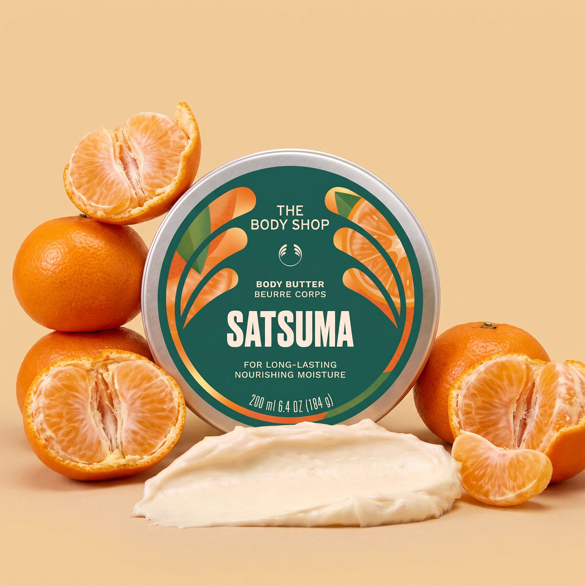 A round tin of The Body Shop Satsuma body butter is centered on a warm beige background, surrounded by whole and sliced oranges, with a smear of creamy off-white body butter in front. The label is dark green with orange and white graphics and text.