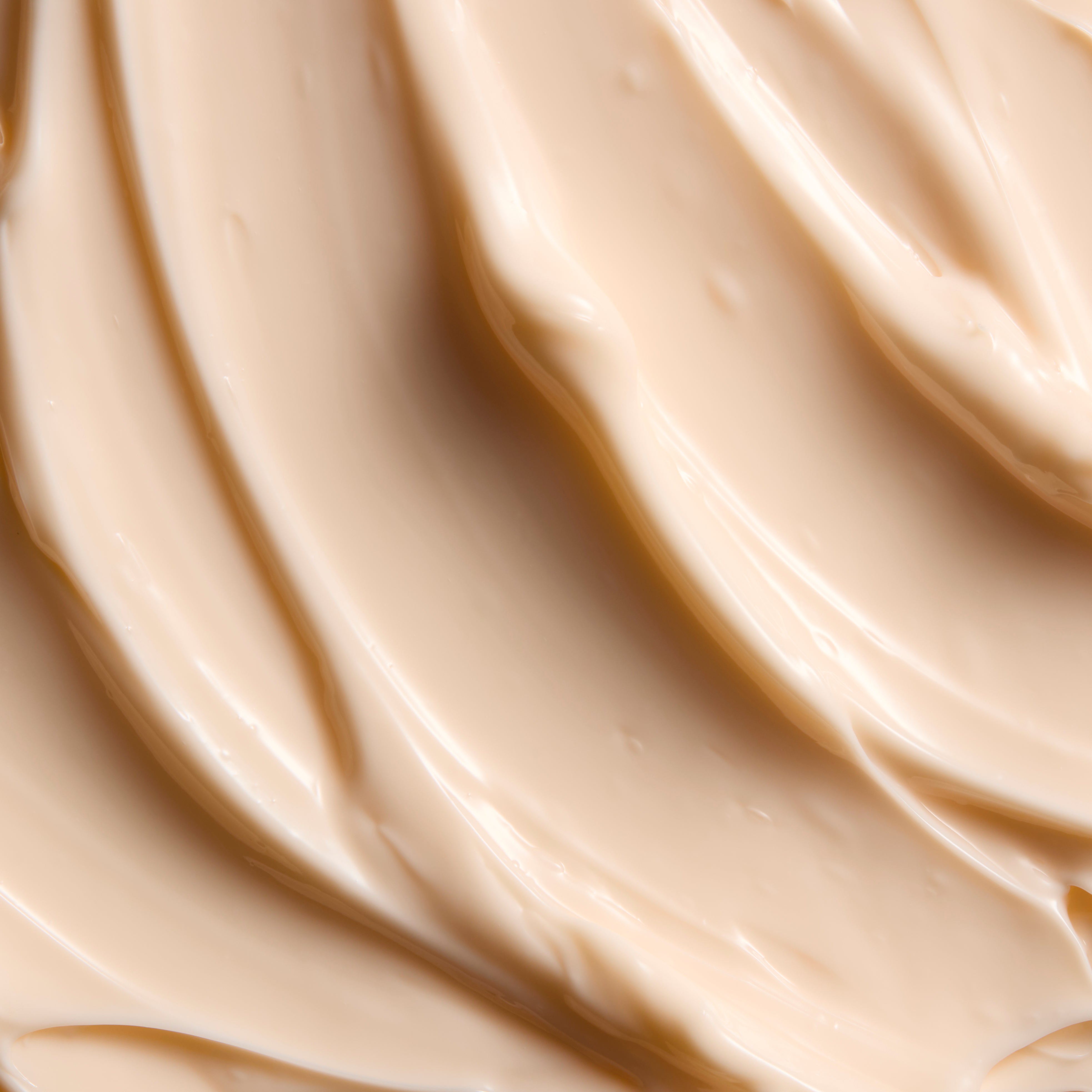 A close-up texture shot of a smooth, creamy beige cosmetic cream with elegant swirled ridges and soft highlights.