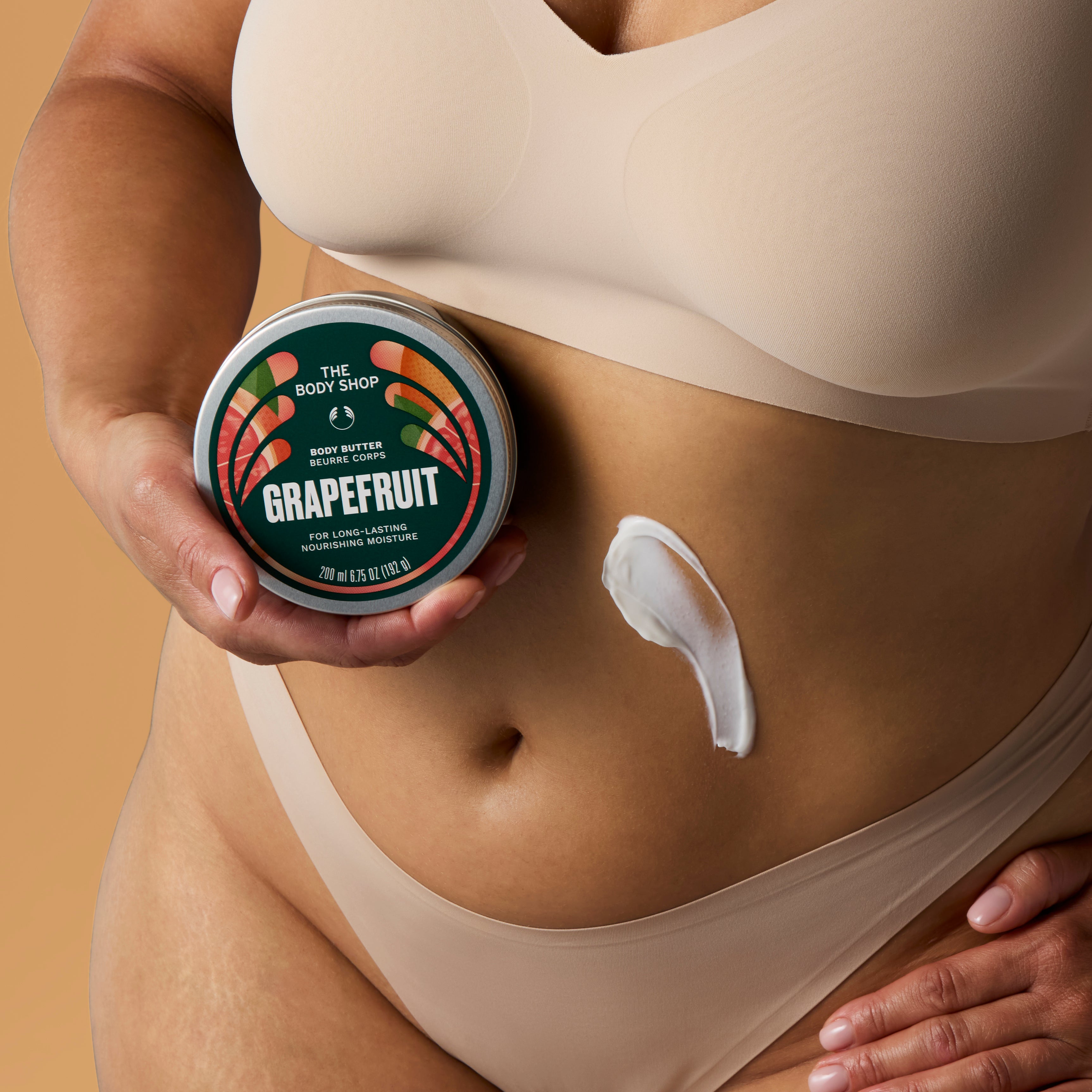 A close-up of a person wearing beige underwear and holding a round tin of The Body Shop Grapefruit Body Butter, with a smear of white body butter on their abdomen against a warm beige background.
