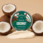 A round tin of coconut body butter by The Body Shop is centered on a beige background, surrounded by whole and halved coconuts with a smear of creamy white body butter in front. The tin label is teal and white with coconut imagery.