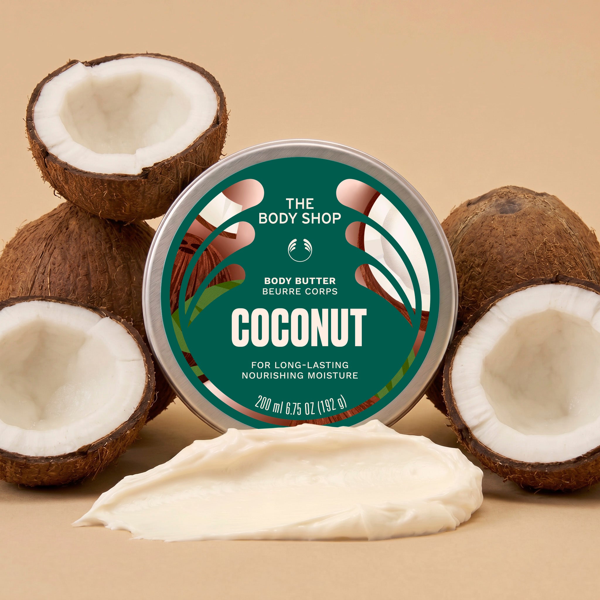 A round tin of coconut body butter by The Body Shop is centered on a beige background, surrounded by whole and halved coconuts with a smear of creamy white body butter in front. The tin label is teal and white with coconut imagery.