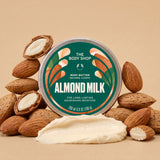 A round tin of The Body Shop Almond Milk Body Butter is centered on a beige background, surrounded by whole and halved almonds with a dollop of creamy off-white body butter in front. The tin label is green and white with almond illustrations.