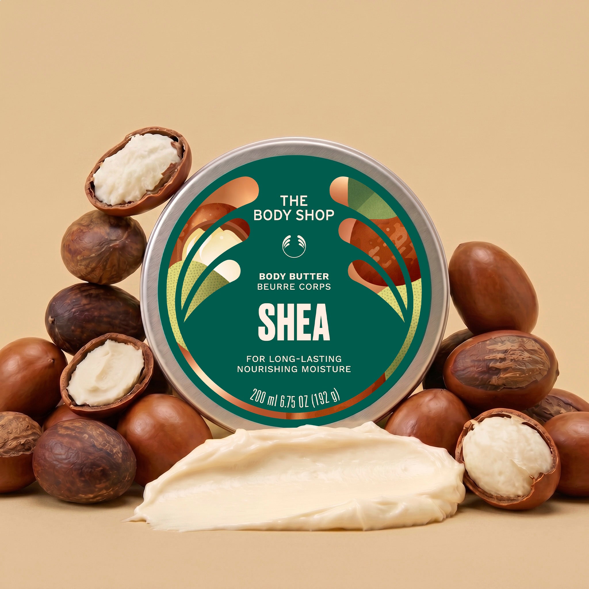 A round tin of The Body Shop Shea Body Butter surrounded by whole and cracked cashews on a warm beige background, with a smear of creamy shea butter in front.