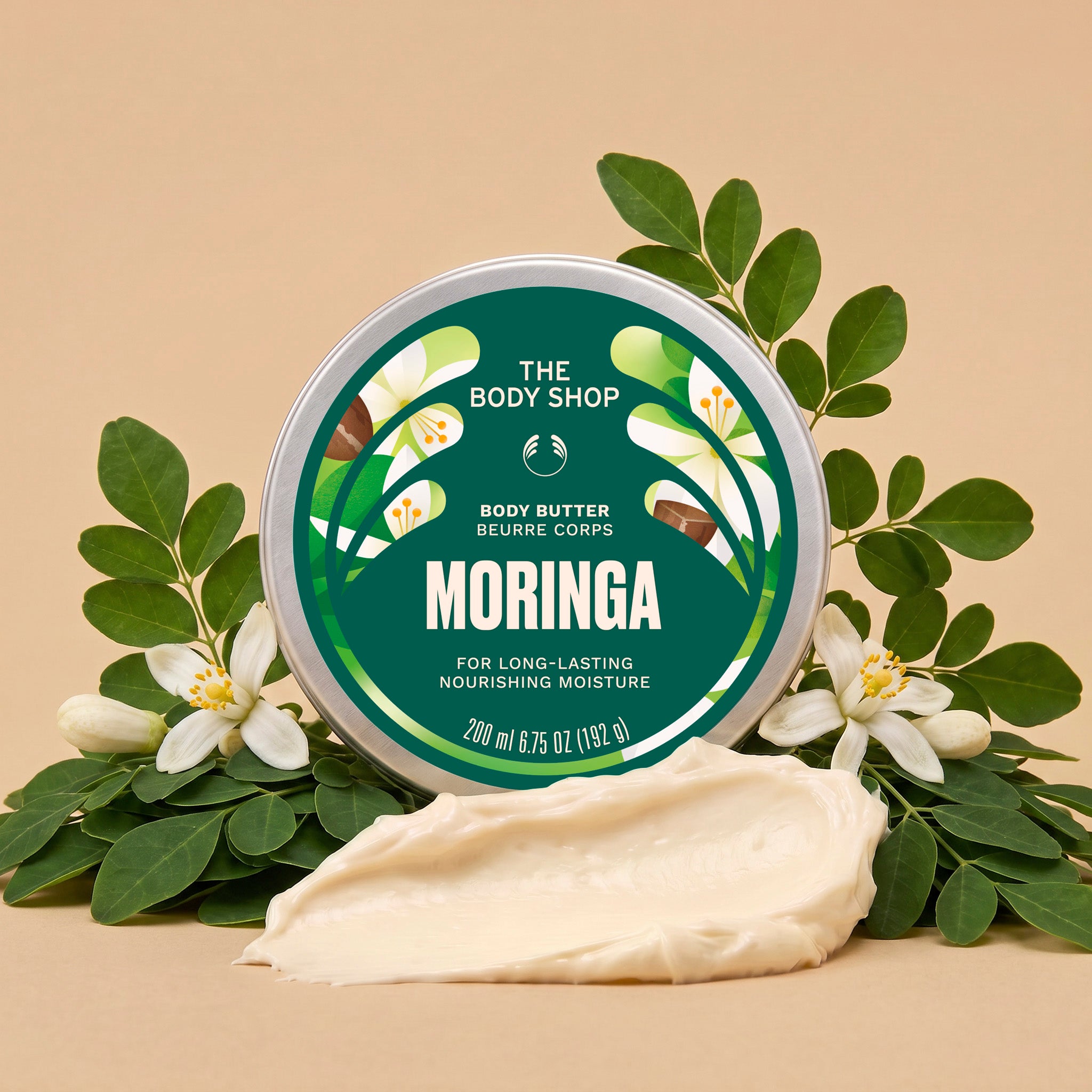 A round tin of The Body Shop Moringa Body Butter sits on a beige surface, surrounded by green moringa leaves and white flowers, with a smear of the cream-colored body butter in front.