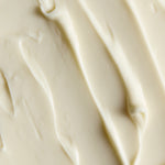 A close-up texture shot of a thick, creamy off-white cosmetic or food product, showing smooth swirled strokes and soft shadows.