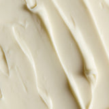 A close-up texture shot of a thick, creamy off-white cosmetic or food product, showing smooth swirled strokes and soft shadows.