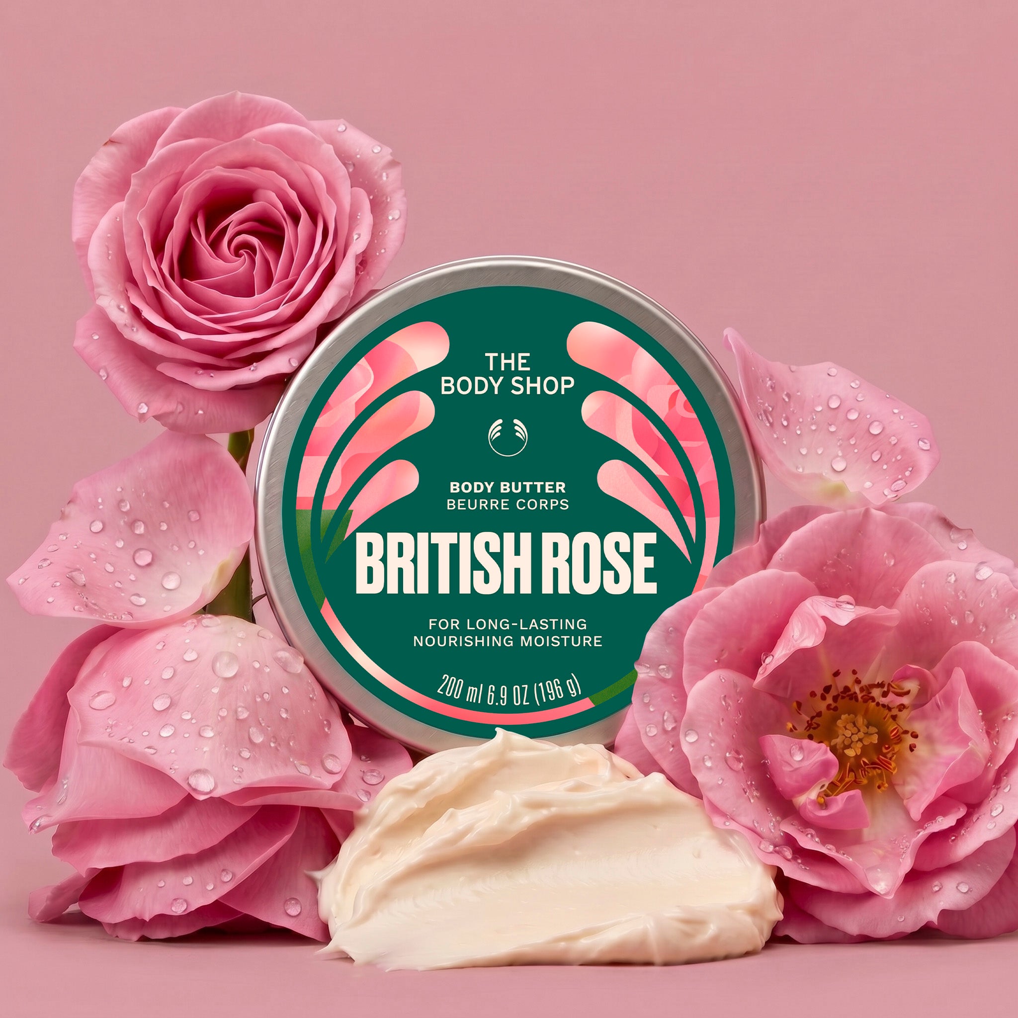 A round tin of The Body Shop British Rose body butter sits on a pink background, surrounded by pink roses and a dollop of creamy off-white body butter. The tin label is dark green with pink rose graphics and white text.