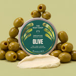 A round tin of The Body Shop Body Butter Beurre Corps Olive moisturizer, surrounded by whole green olives with a smear of olive-colored body butter in front, on a light olive-green background.