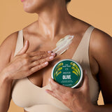 A woman in a beige bra holds a round tin of The Body Shop Olive Body Butter & Mask against a warm beige background, with cream-colored body butter smeared on her chest.