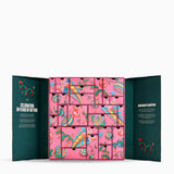 An open advent calendar with a pink front panel featuring many small illustrated drawers, flanked by dark teal inner flaps with decorative text and red botanical details, shown on a light gray background.