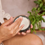 Person applying a white cream from a jar to their arm with a blurred indoor background