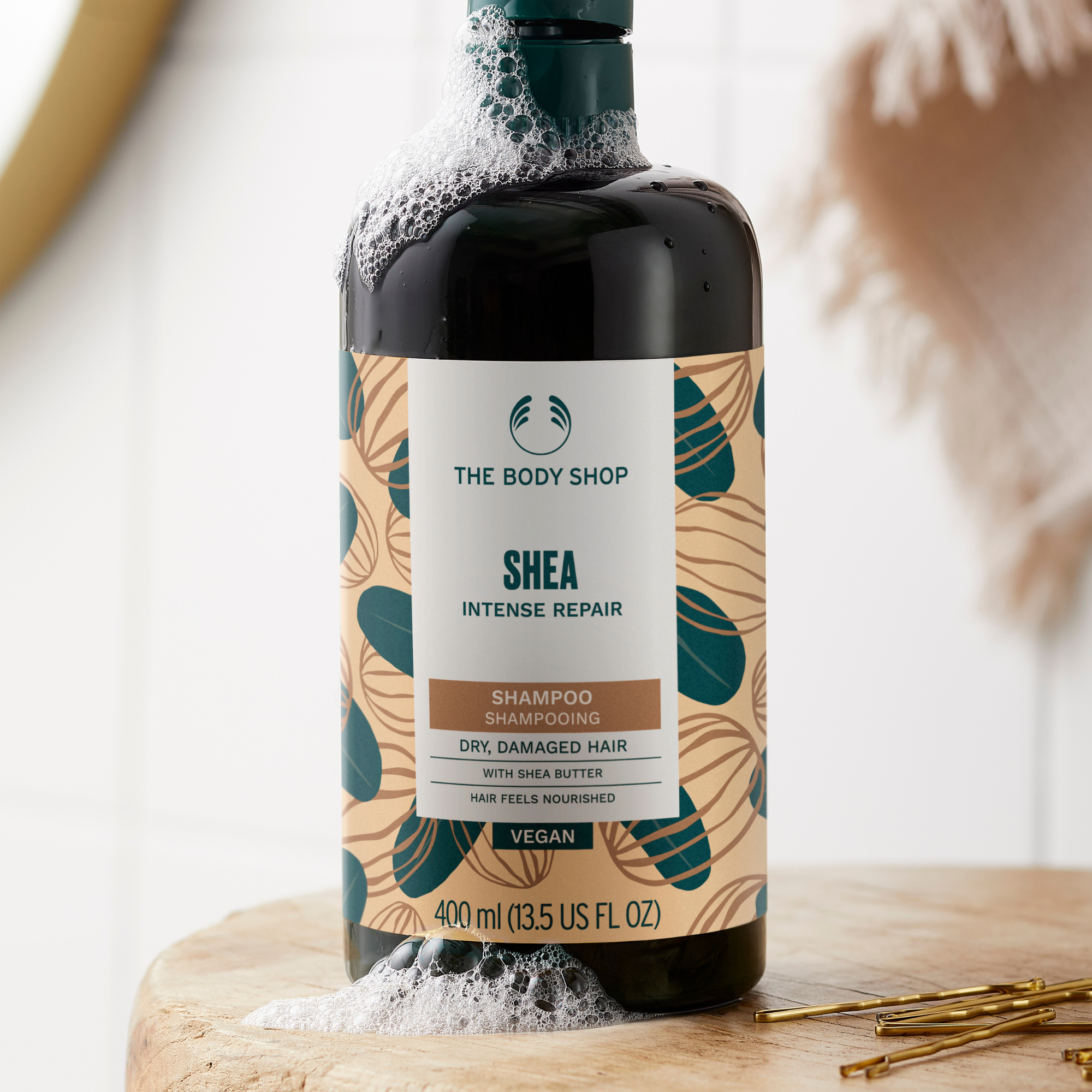 A bottle of The Body Shop Shea Intense Repair Shampoo standing on a wooden surface with white foam around the base. The bottle is dark with a patterned beige label featuring teal and brown botanical motifs.