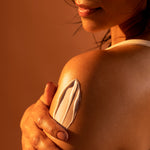 Person applying body cream to shoulder with a warm brown background