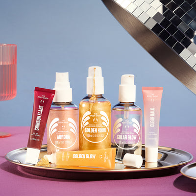 Four COSRX skincare products from the Clear To Glow line are arranged on a silver tray against a blue and purple studio background. The products include a Crimson Flare tube, an Aurora moisturizer bottle, a Golden Hour moisturizer bottle, a Solar Glow spray, and a Clear Halo tube, with a Golden Glow bar in front.