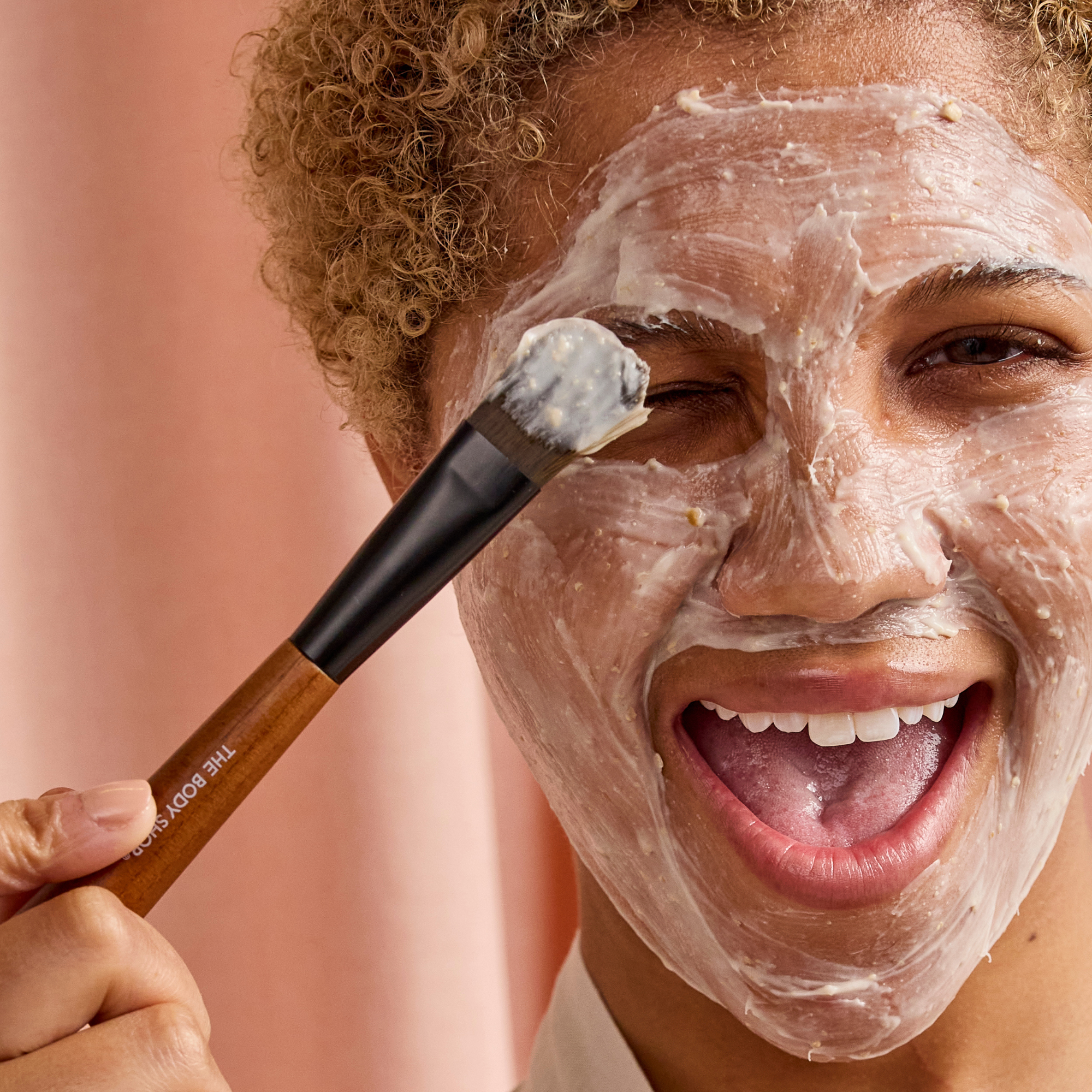 Person applying a facial mask with a brush on a pink background