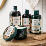 Four The Body Shop Shea Intense Repair hair care products arranged on a wooden surface: two dark shampoo bottles, one shampoo bottle with its lid off showing white shampoo inside, and one dark conditioner jar. A wooden comb rests beside them against a bright tiled background.