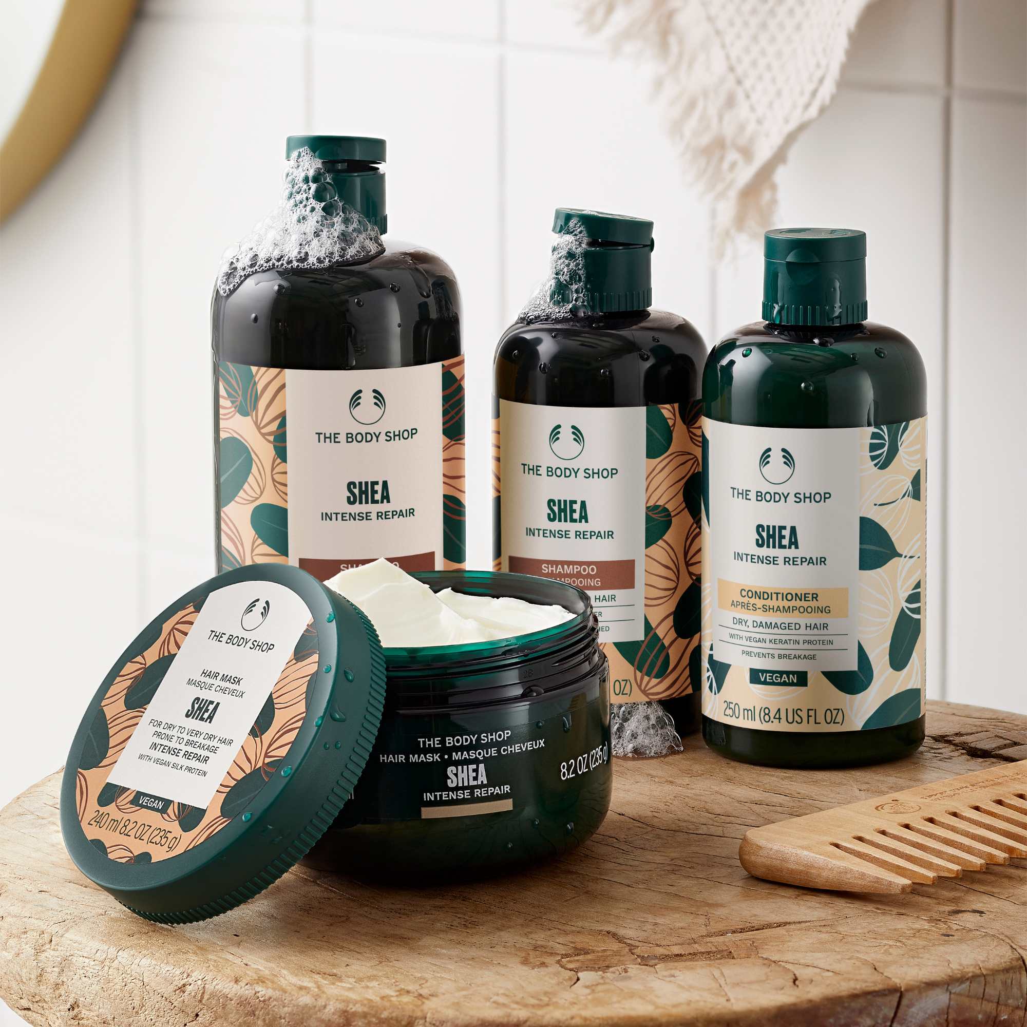Four The Body Shop Shea Intense Repair hair care products arranged on a wooden surface: two dark shampoo bottles, one shampoo bottle with its lid off showing white shampoo inside, and one dark conditioner jar. A wooden comb rests beside them against a bright tiled background.