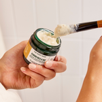 Hand applying cream from a jar labeled 'The Body Shop' with a brush.
