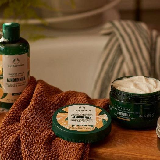 The Body Shop Almond Milk products on a wooden surface with a towel and bathtub in the background.