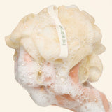 A close-up of a hand lathering soap with a beige, textured scrubber or loofah-like cleansing tool covered in foam. A white loop with green text reading 'THE BODY SHOP' is visible on the tool.