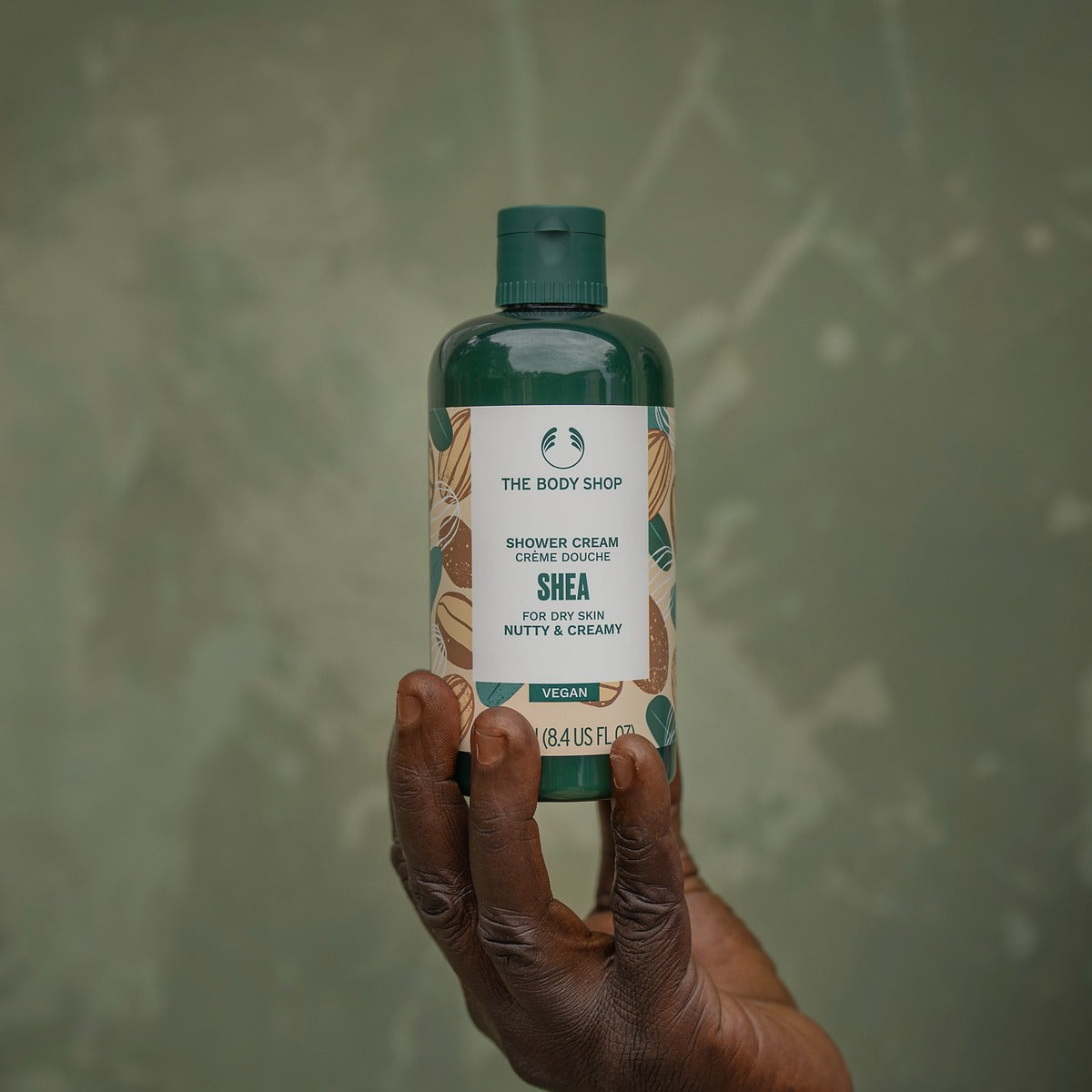 Hand holding a bottle of The Body Shop Shea Shower Cream against a green background