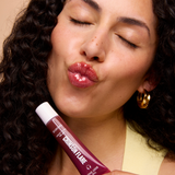 A close-up lifestyle shot of a woman with curly dark hair and closed eyes, puckering her lips to show glossy pink lips, while holding a tube of Chamilia Flare lip gloss near her face.