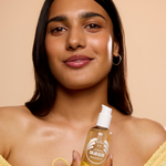 A woman with a radiant complexion holds a bottle of Solar Glow hair oil against a warm beige background. The bottle is transparent, showing the golden-yellow oil inside, and features a white pump dispenser.