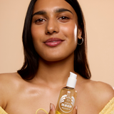 A woman with a radiant complexion holds a bottle of Solar Glow hair oil against a warm beige background. The bottle is transparent, showing the golden-yellow oil inside, and features a white pump dispenser.