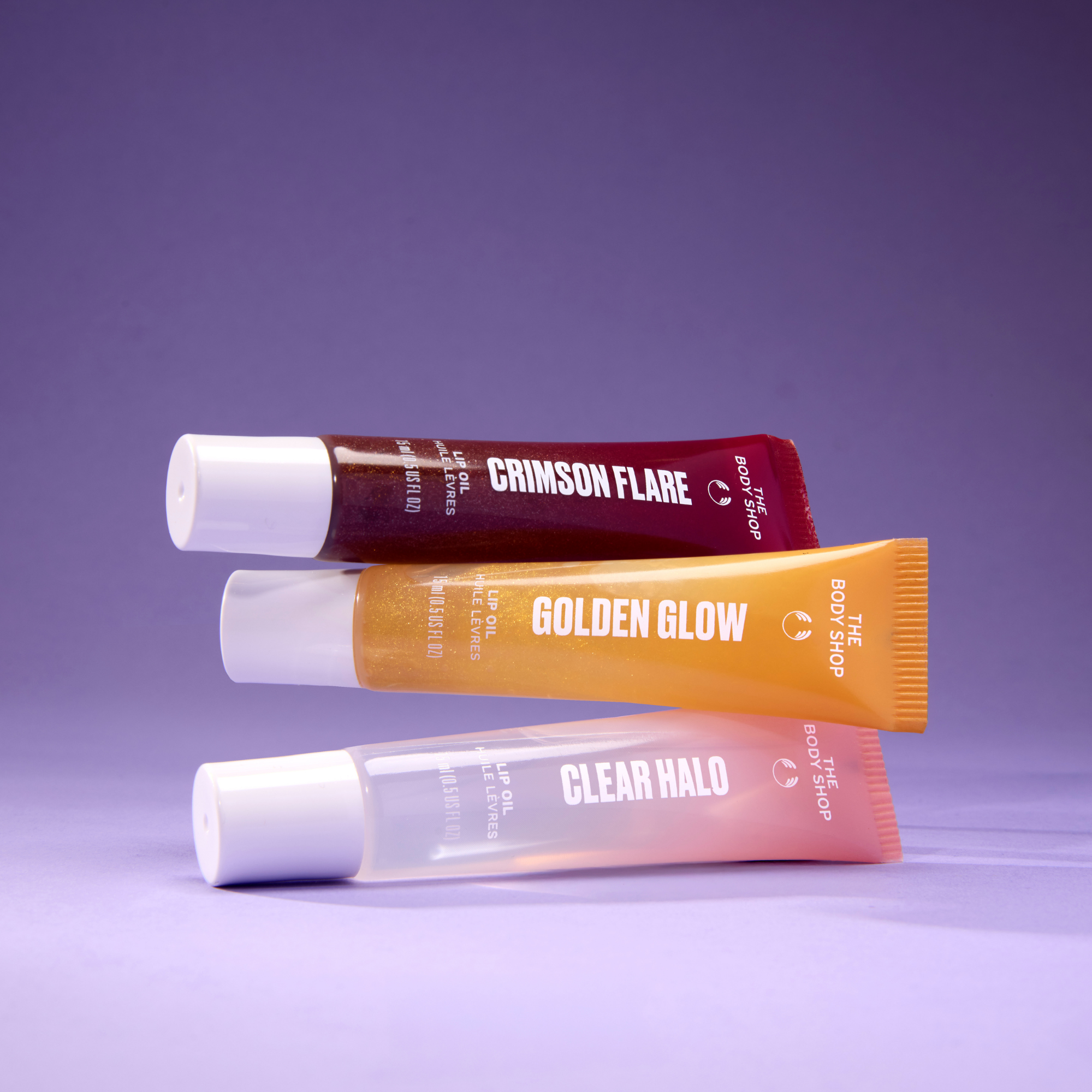 Three lip balm tubes from The Body Shop's 'The Lip Shop' are stacked diagonally on a purple background. The tubes are labeled 'CRIMSON FLARE' in dark red, 'GOLDEN GLOW' in orange, and 'CLEAR HALO' in translucent pink, all with white caps.