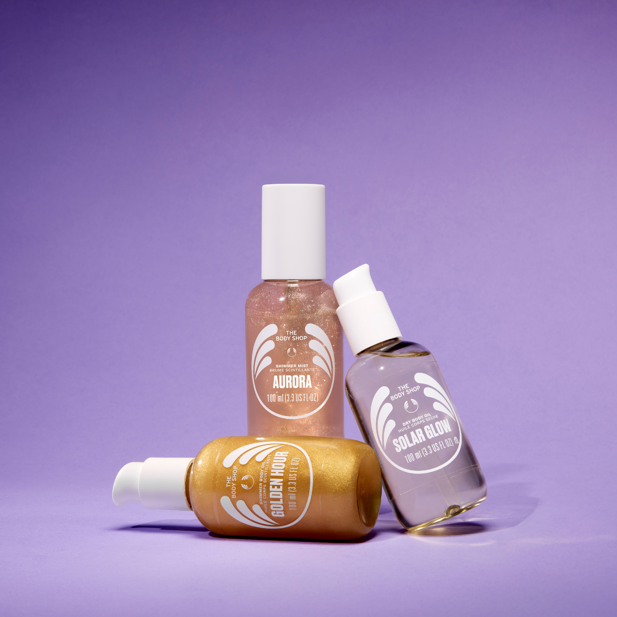 Three cosmetic bottles from The Art of Beauty arranged on a solid purple background. The set includes a gold pump bottle labeled Golden Hour, a clear bottle with white cap labeled Aurora, and a clear spray bottle labeled Solar Glow.
