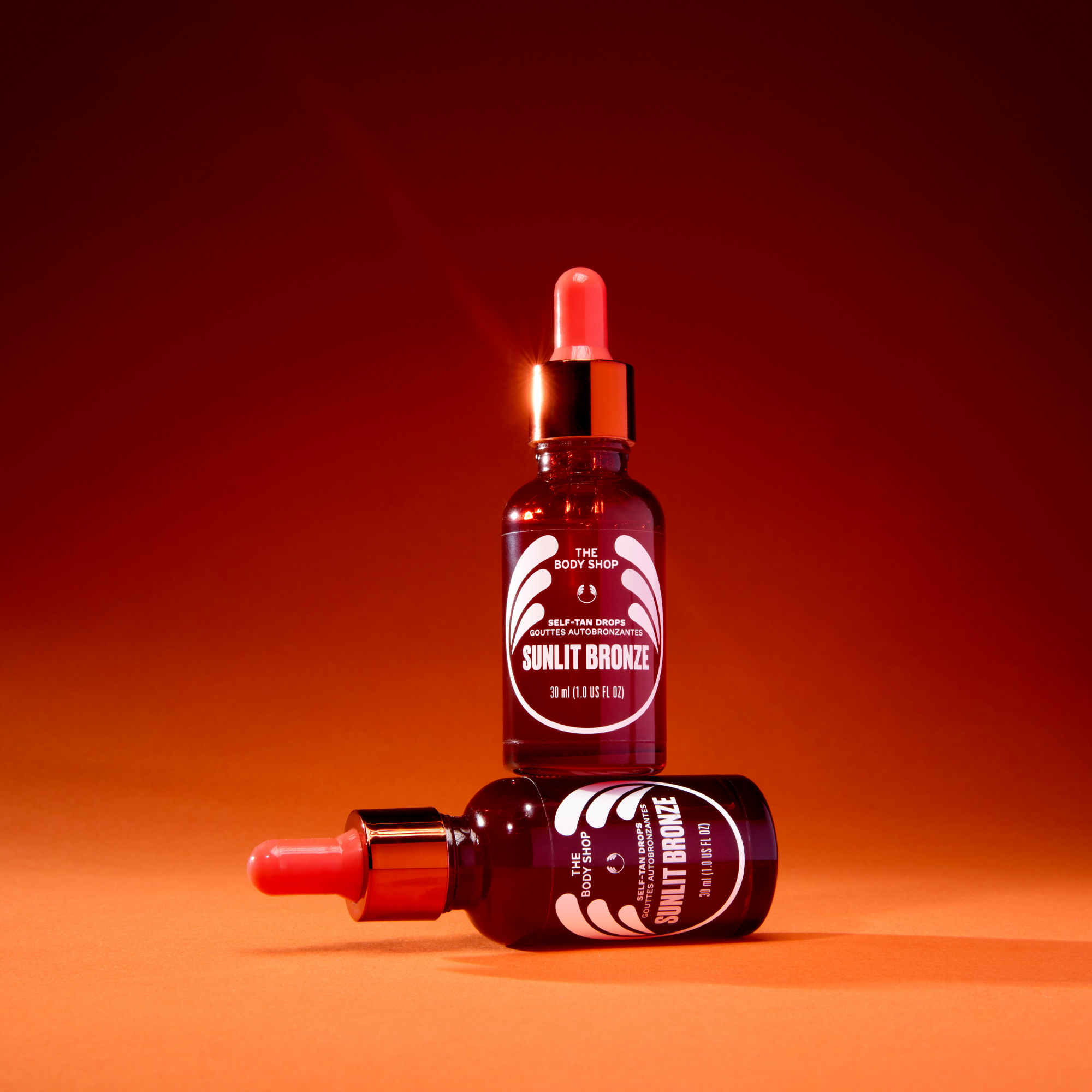Two amber glass dropper bottles of The Body Shop Sunlit Bronze hair color, one standing upright and one lying on its side, set against a warm red-to-orange gradient background.